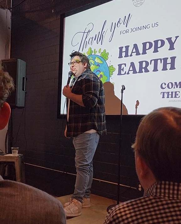 A stand-up comedian performing on stage at an event titled 'Happy Earth' with a large screen in the background displaying the event name and a graphic of the Earth.