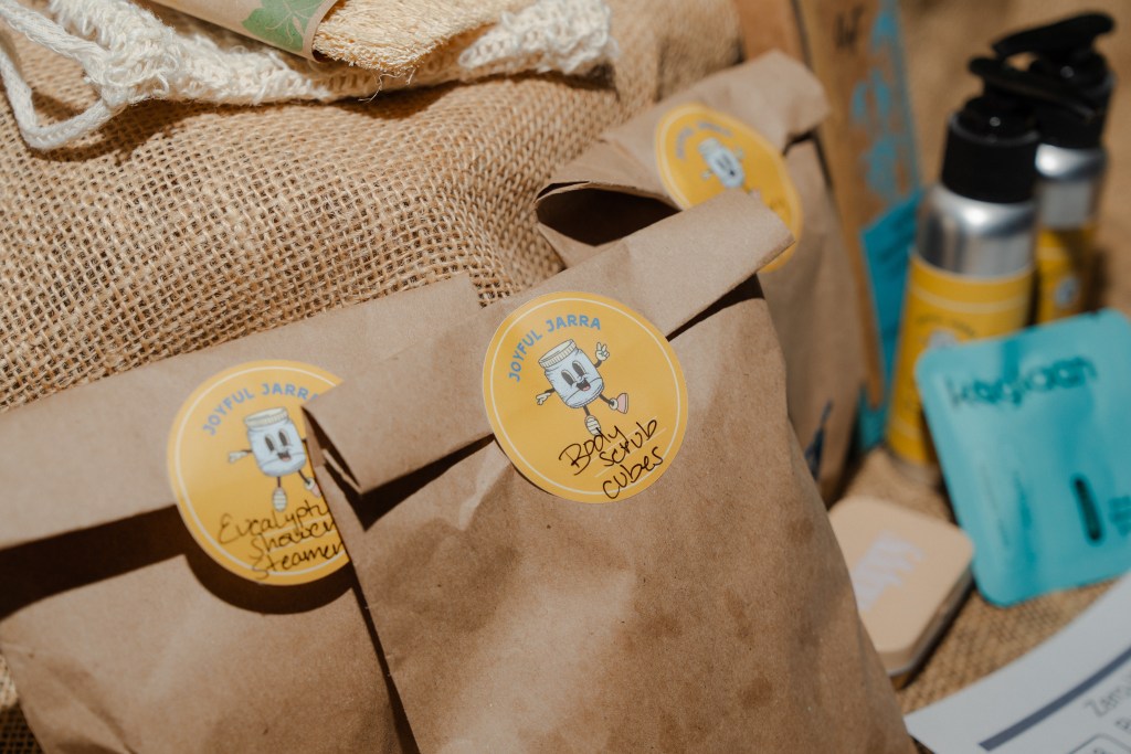 Brown paper bags labeled 'Eucalyptus Shower Steamer' and 'Body Scrub Cubes' on a textured burlap background, surrounded by various skincare products.