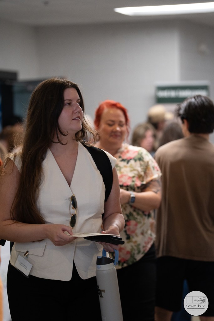 A woman with long hair holding a notebook stands in a crowded hallway, wearing a white sleeveless top and sunglasses. In the background, several people are mingling, with one person having red hair and wearing a floral shirt.