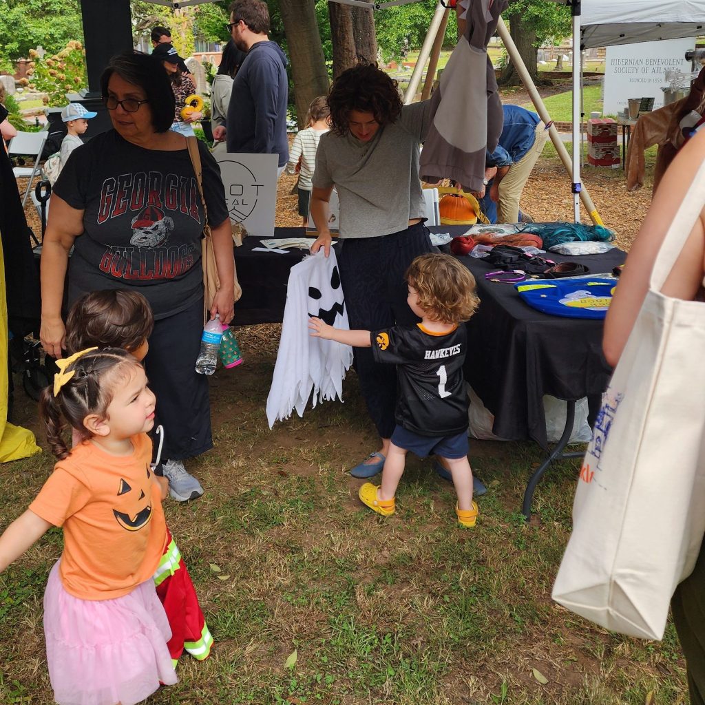 Children playing near a craft booth at an outdoor event, with adults supervising. One child is dressed in a pumpkin-themed shirt and colorful skirt, while another child holds a white ghost costume. Various activities and colorful items are visible on the table.